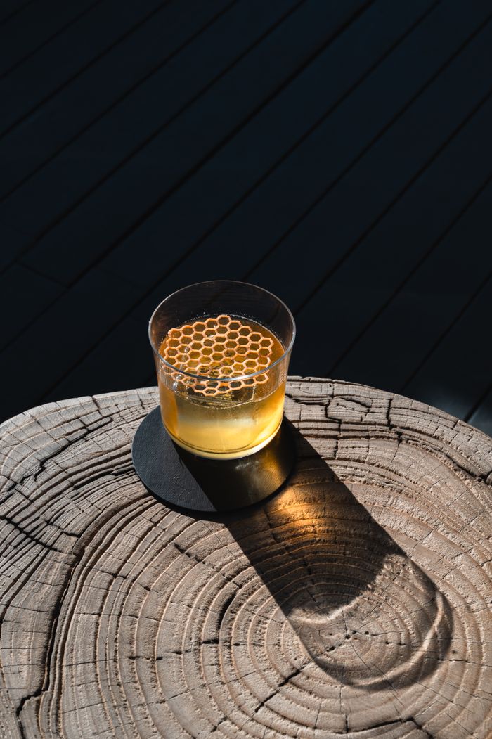 Whiskey cocktail with honeycomb on a wooden stump under harsh lighting.