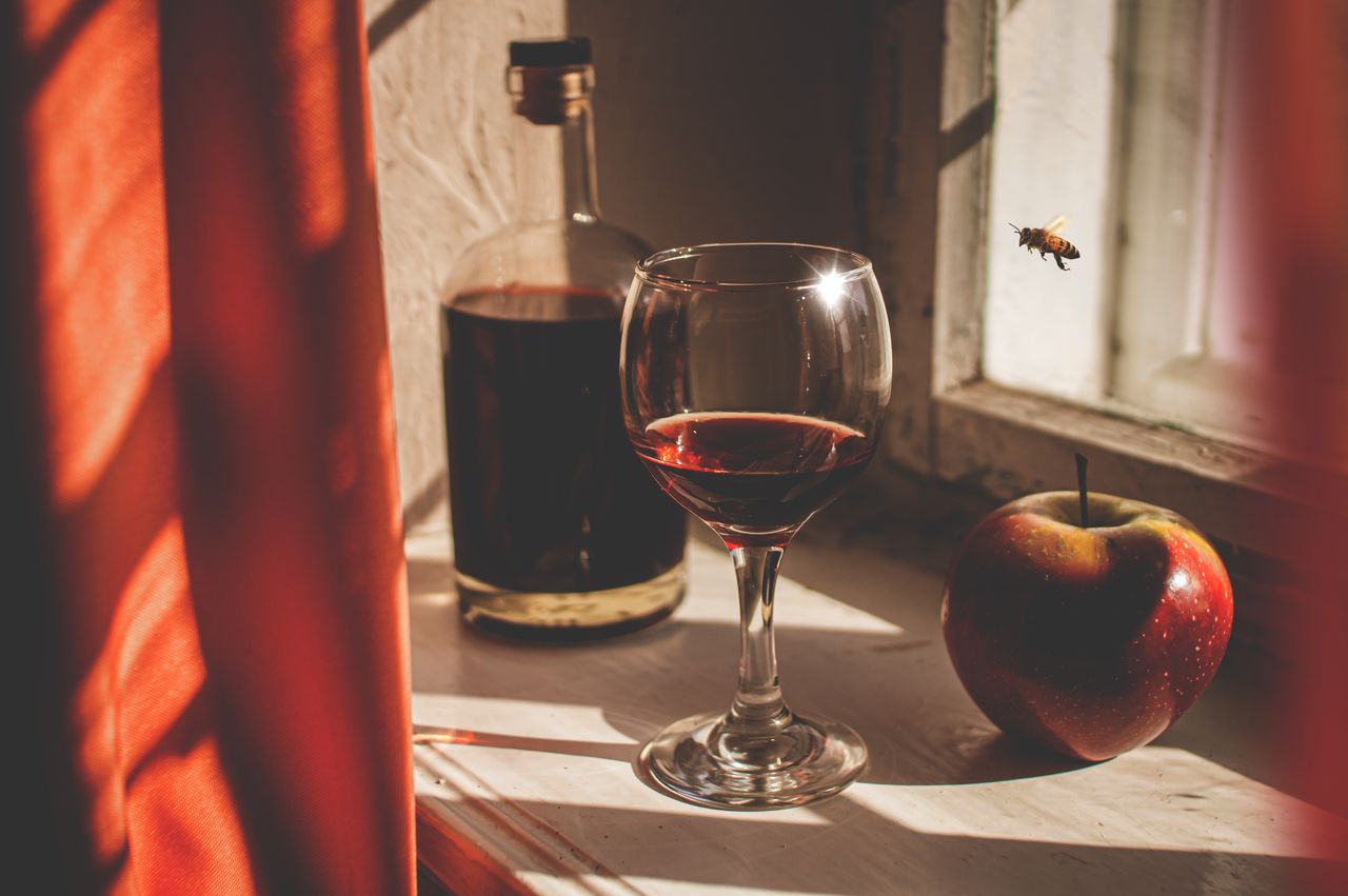 About A glass of red wine, apple, and bottle on a sunlit windowsill with a bee hovering.